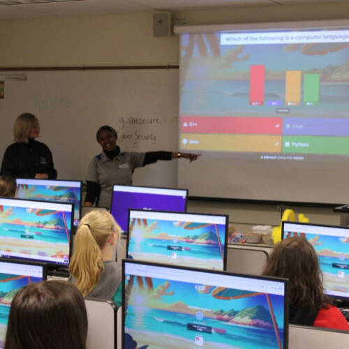 A classroom of students at desktop computers watches as two instructors lead an interactive quiz about computer languages, displayed on a projector at the front of the room.